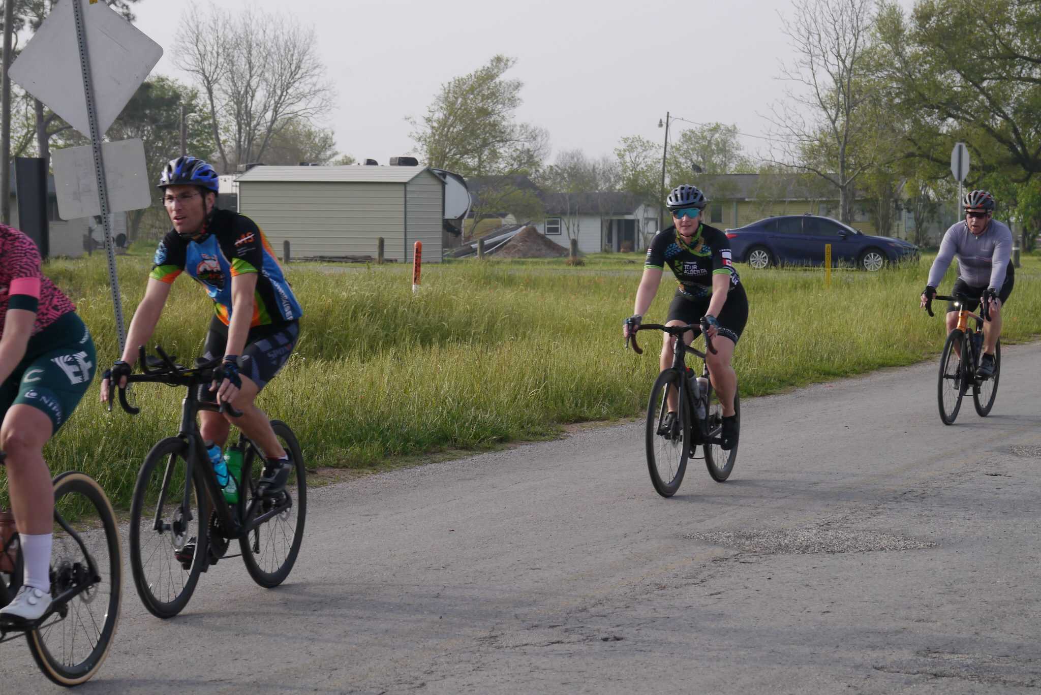 Fast cyclists riding on a rural road with grass and trees in the background, promoting Bluebonnet Express bike rides.