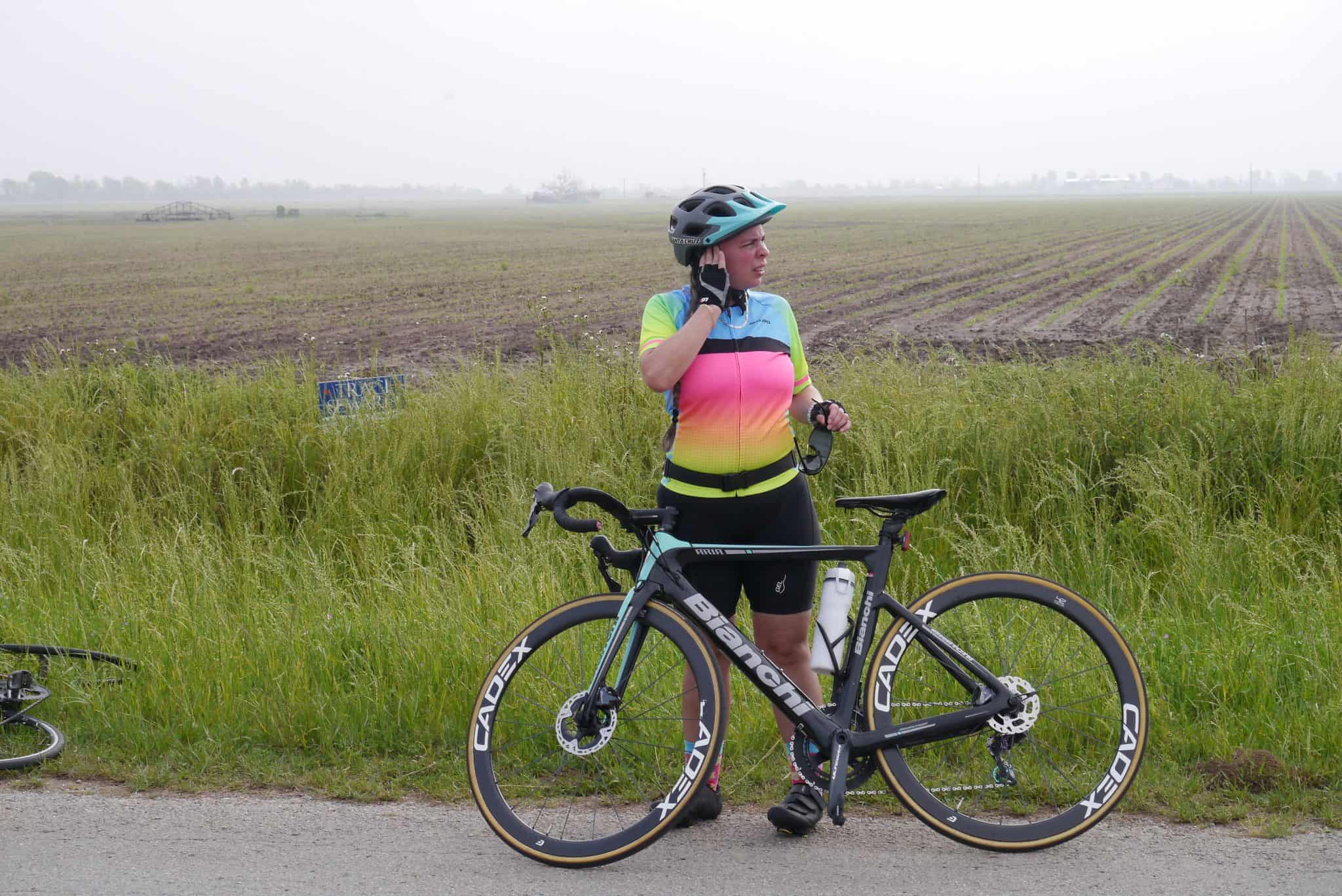 Bright female cyclist with a Bianchi road bike on rural roadside, wearing colorful gear and helmet.
