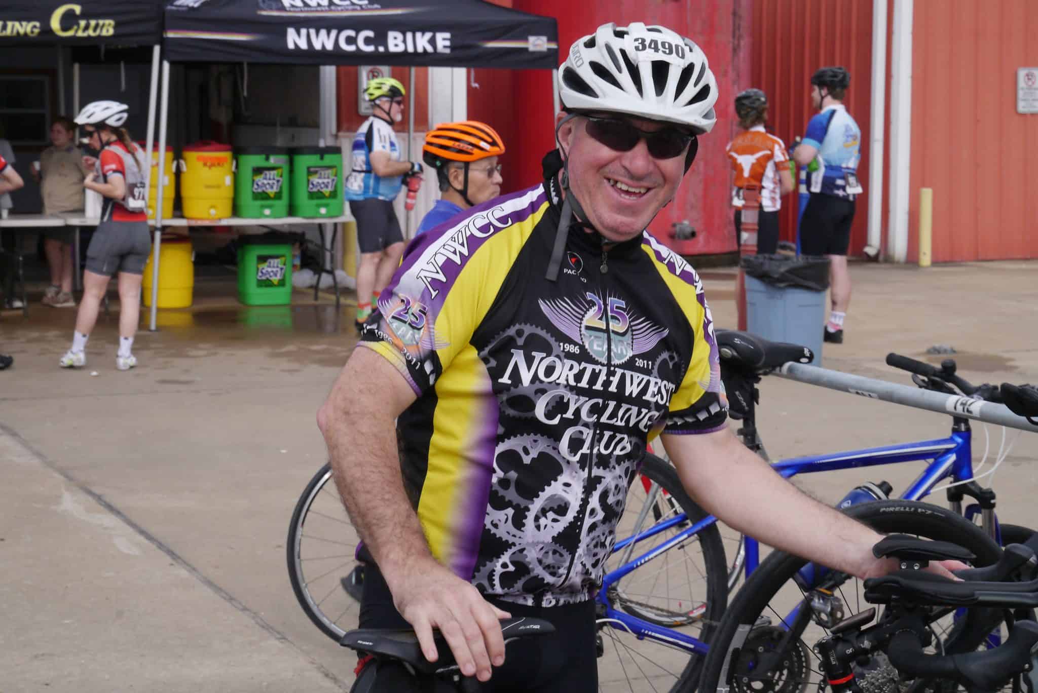 Smiling cyclist in Northwest Cycling Club jersey at Bluebonnet Express event, with bikes and fellow riders in the background.