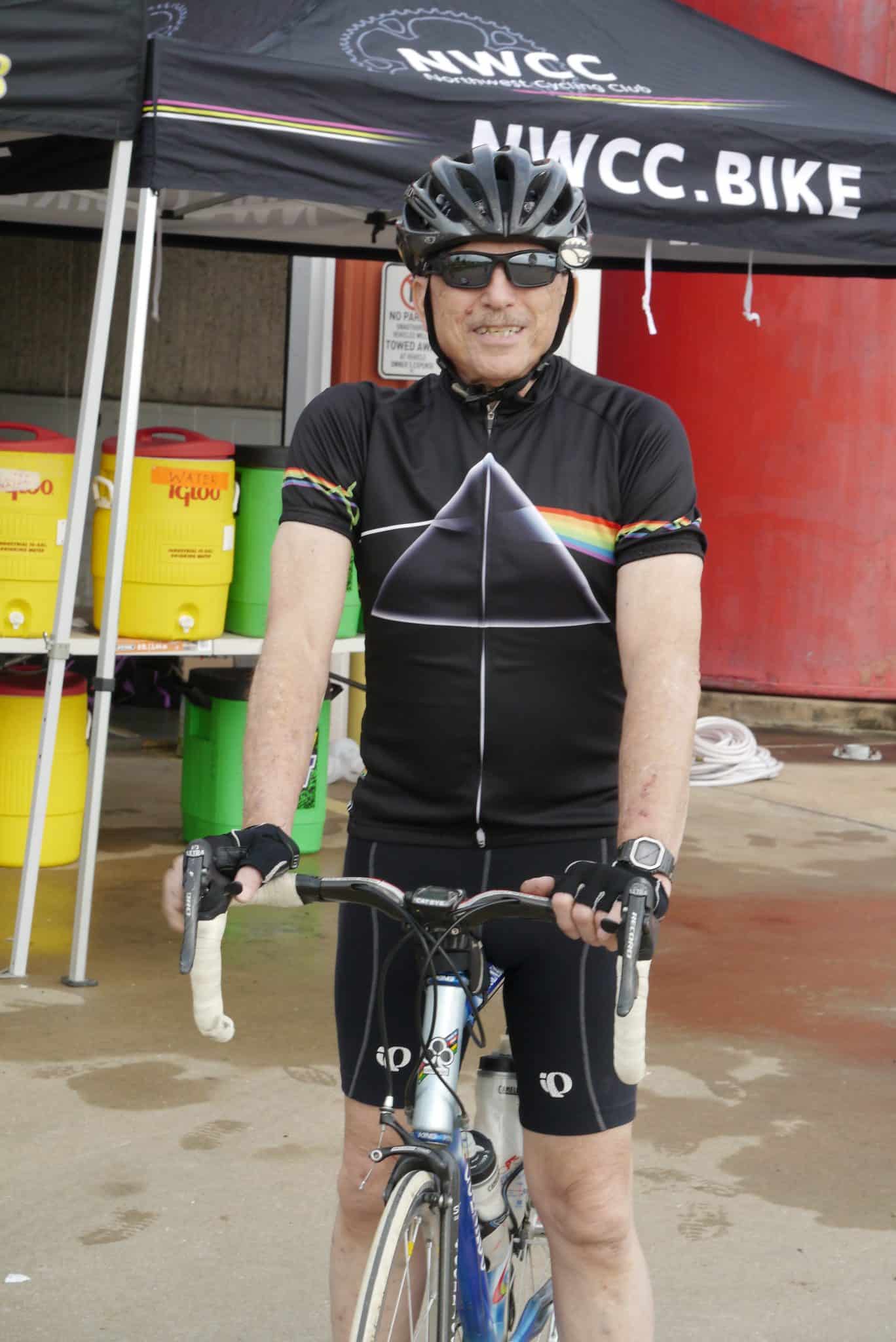 A man in cycling gear with a Black helmet and sunglasses standing with a bicycle outdoors.