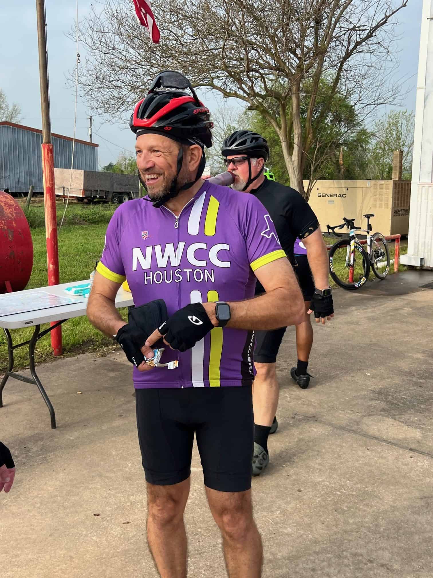 Smiling cyclist in purple NWCC Houston jersey, ready for a bike ride with friends, outdoor setting, trees, bikes in background.