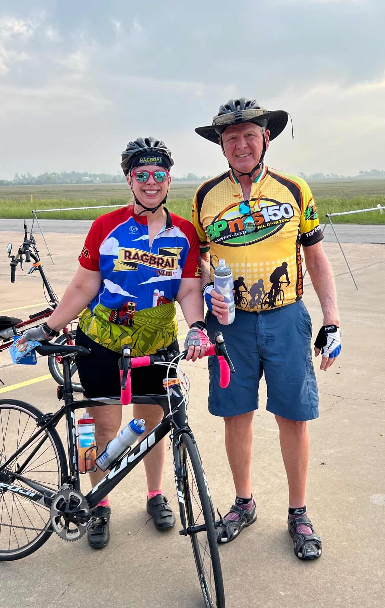 Bright female cyclist with mountain bike and male cyclist in race jerseys outdoors against a cloudy sky.