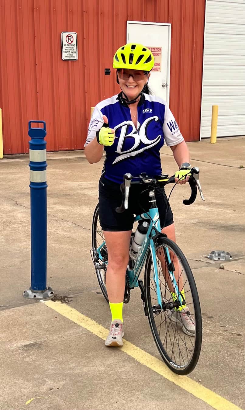 Bright female cyclist wearing Bluebonnet Express jersey with cycling gear, standing with her bike outdoors.