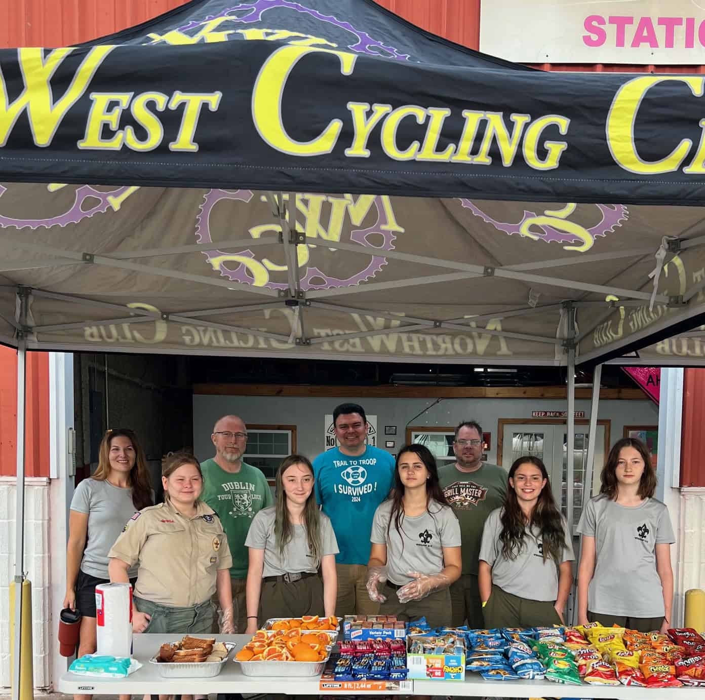 Bike shop booth at cycling event, people volunteering and selling snacks, Promotional cycling gear at West Cycling Club.