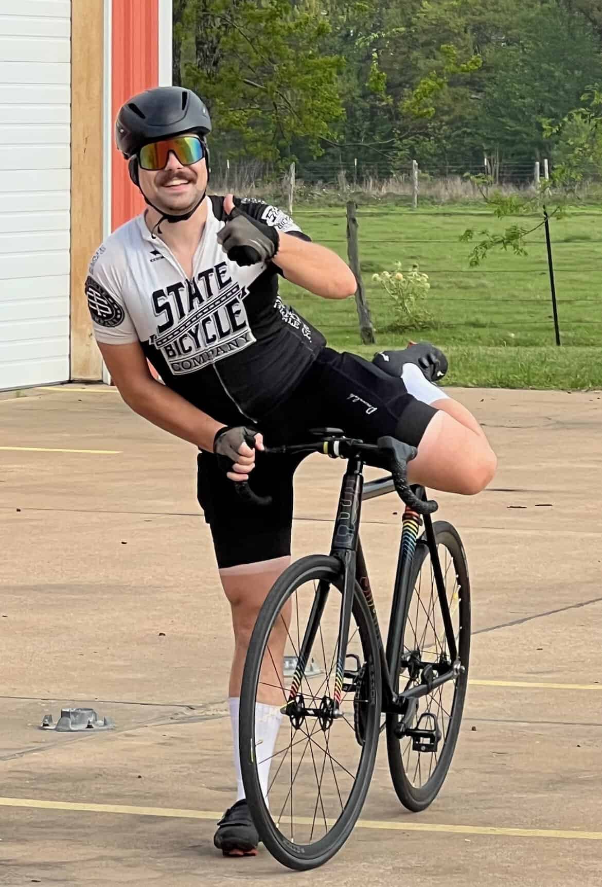 Cyclist with a Black Road Bike, wearing a helmet and sunglasses, smiling, celebrating outdoors.